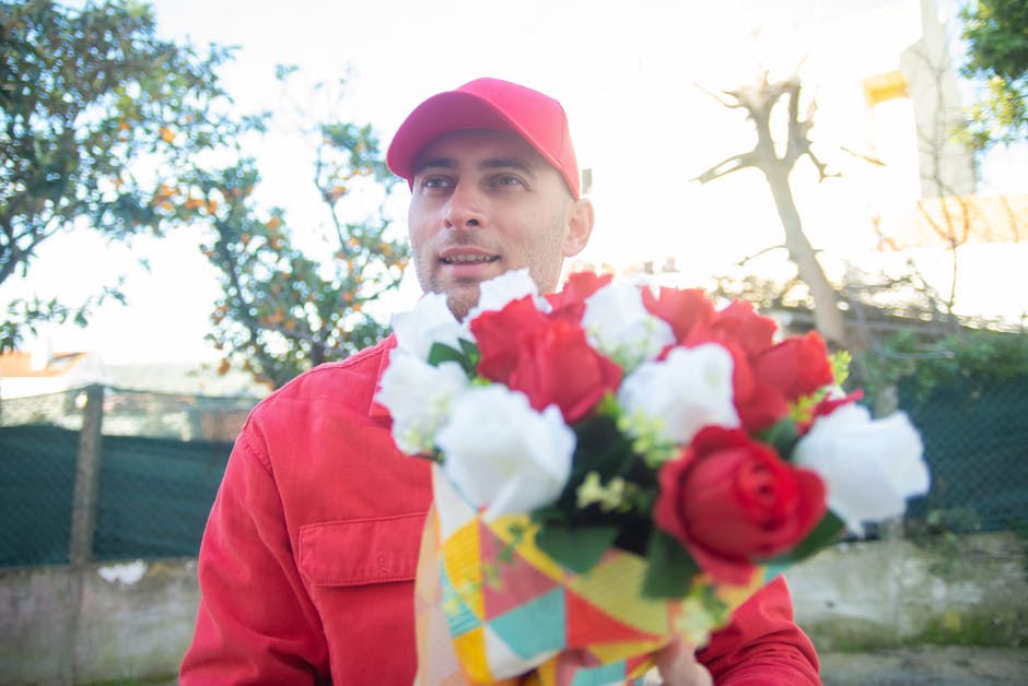 A man wearing a red cap and matching red jacket is holding a vibrant bouquet of fresh flowers, including red roses and white blossoms, wrapped in colorful paper. The bouquet is positioned in front of