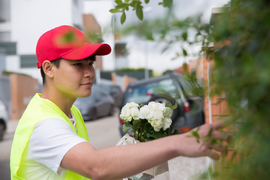 A young delivery person wearing a red cap and a neon yellow vest hands over a bouquet of fresh, white hydrangeas wrapped in kraft paper to a recipient at an outdoor setting. The flowers are vibrant an