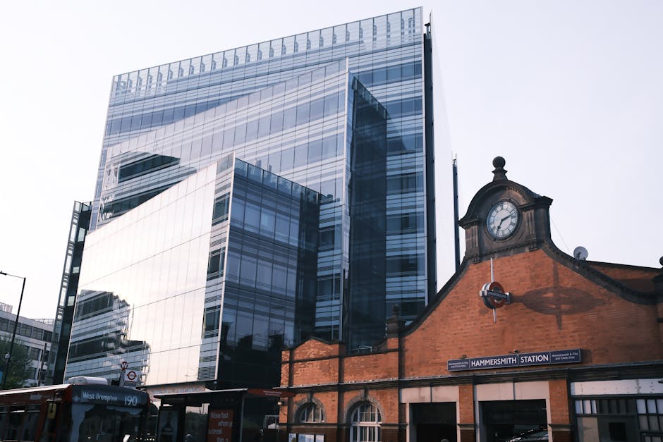 A modern glass office building reflecting the sky and surrounding structures stands behind the historic brick Hammersmith Station building, which features a decorative clock tower and a station sign. 