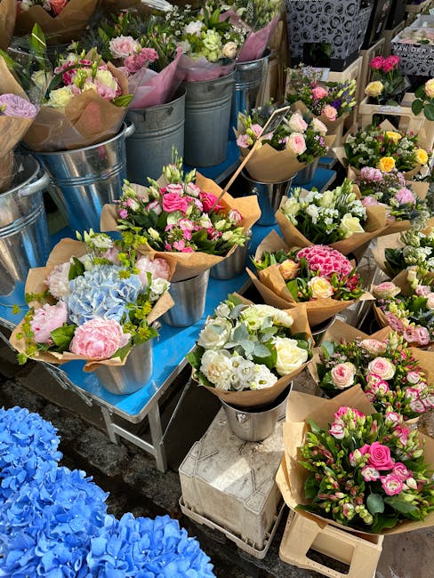 An assortment of fresh flowers arranged in small bouquets and wrapped in brown kraft paper, displayed in metal buckets and vases on a blue table at a flower market. The bouquets feature pastel-colored