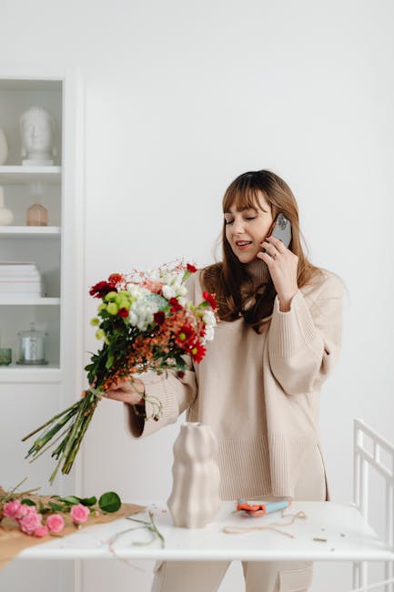 A woman with long brown hair and a beige sweater is holding a vibrant bouquet of fresh flowers, including red, white, and pink blossoms, with green foliage, in her left hand. She is speaking on a mobi