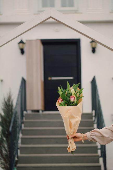 A person holding a bouquet of fresh flowers featuring pink tulips and green foliage wrapped in light brown paper, positioned in front of a modern white house with a dark door. The bouquet displays vib
