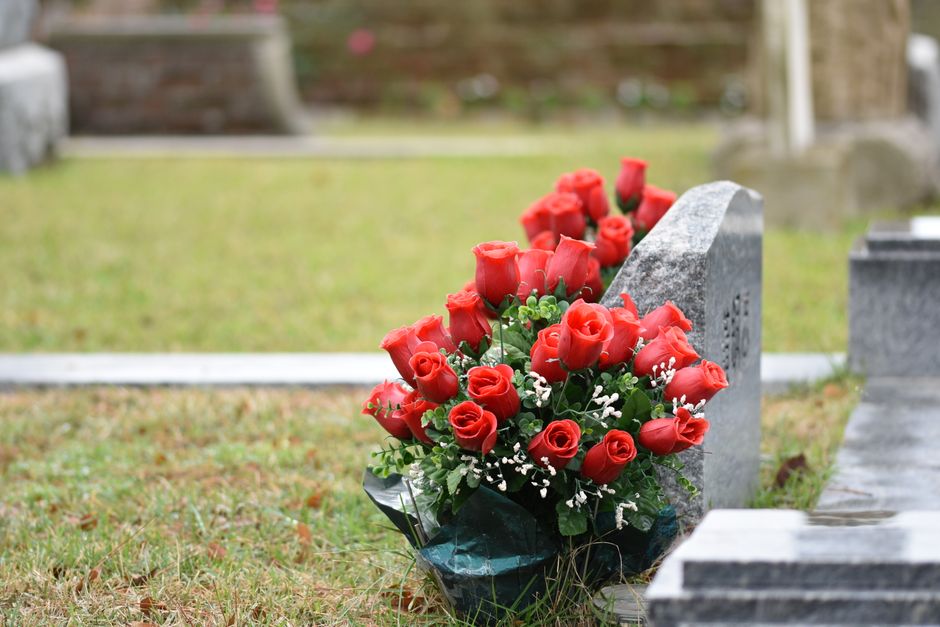 A bouquet of fresh red roses with green foliage and small white filler flowers, arranged in a rounded, tightly clustered style, rests against a gray gravestone in a cemetery. The roses have vibrant, v