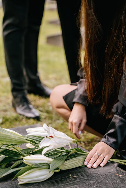 A woman with long brown hair is kneeling on the ground, gently placing her hand on a floral arrangement comprised of white lilies with large petals and prominent stamens, and lush green leaves. She is