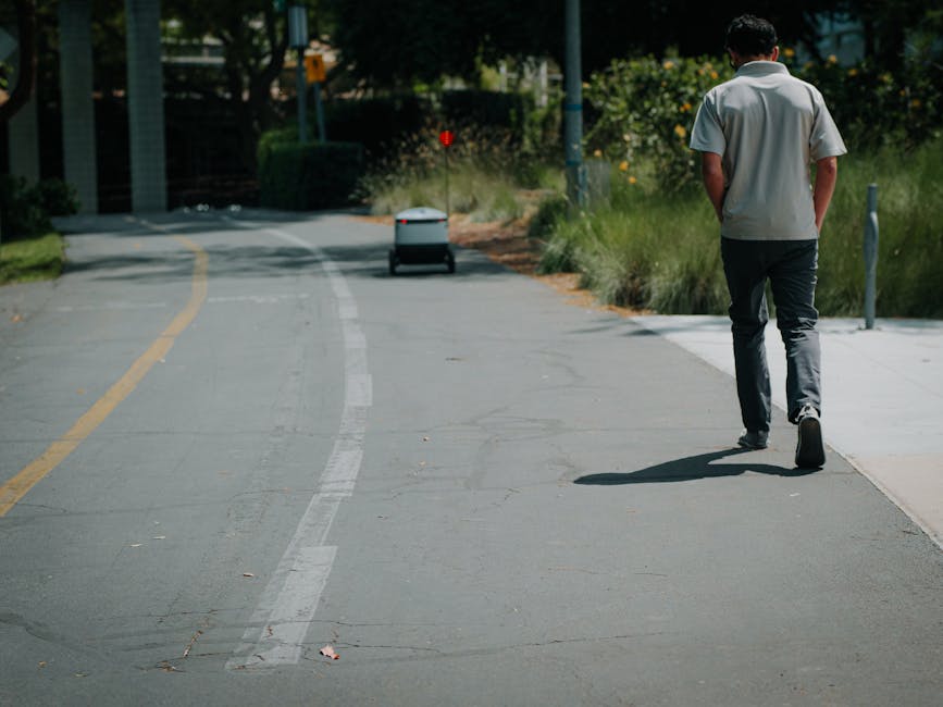A man dressed in a light grey shirt and dark trousers walking along a paved sidewalk next to a bicycle lane, with lush green plants and trees on the side. The background features a small autonomous de